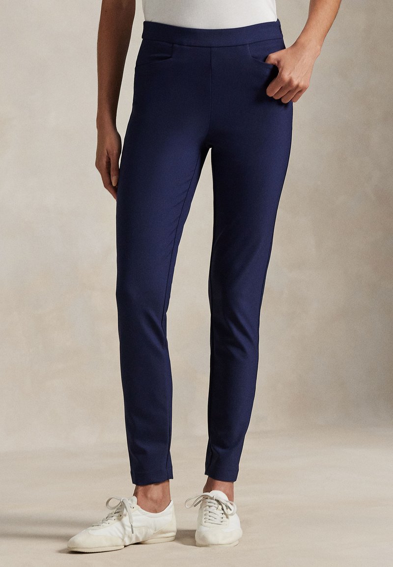 Persoon die slim-fit marineblauwe broek en witte sneakers draagt, staand tegen een eenvoudige neutrale achtergrond met één hand in de zak.