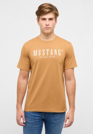 Young man with light brown hair wears a mustard yellow t-shirt with "MUSTANG Jeans Company Since 1932" text and blue jeans, standing against white background.