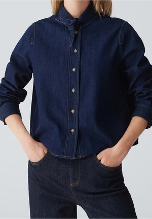 Person wearing dark blue denim shirt with button-up front and matching dark denim jeans, hands in pockets, standing against plain background.
