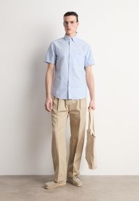 Camisa de botão de manga curta azul clara combinada com calças largas bege e sapatos bege de slip-on, segurando uma mala tote clara.