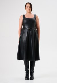 Zwarte faux leren jurk met vierkante halslijn, brede bandjes en ceintuur in de taille. Bevat een vloeiende rok en zwarte kniehoog boots.