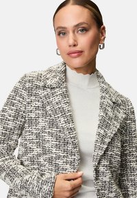 Gestructureerde zwart-wit blazer met een klassiek reversontwerp; heeft zichtbare stiksels en een open voorkant. Draagt over een lichtgrijze top.