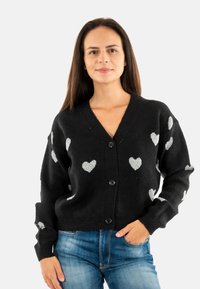 Cardigan nero con motivi a cuore argentati, scollo a V, chiusura con bottoni, polsini e orlo a costine, texture morbida. Indossato sopra jeans blu.