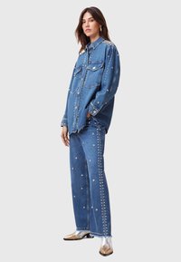 Outfit in denim con una camicia a maniche lunghe e vestibilità ampia con dettagli in borchie e pantaloni a gamba larga, entrambi in blu medio. Tessuto texturizzato con borchie lucide.