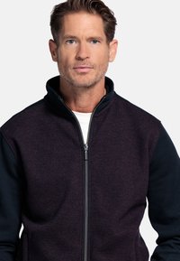 Heren zip-up jack met een bordeauxrood en zwart textuurontwerp, voorzien van een hoge kraag en contrasterende mouwen. Ritssluiting aan de voorkant.