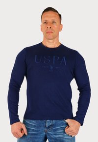 Mornarsko majica s dolgimi rokavi iz bombaža, ki ima na sprednji strani natisnjen logotip v nekoliko temnejši odtenek in okrogel vrat.
