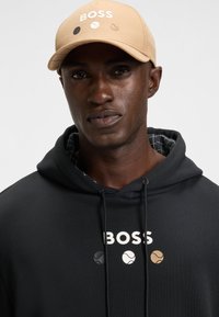 Cappellino da baseball color nocciola con logo "BOSS" e tre design di palline da tennis; indossato con un hoodie nero con un logo bianco e lacci.