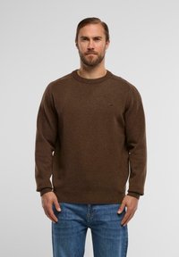 Brauner Strickpullover mit Rundhalsausschnitt, gerippten Bündchen und Saum. Mit dezentem Logodetail auf der Brust. Getragen mit blauen Jeans.
