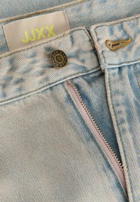 Jean en denim bleu clair avec une fermeture éclair partiellement ouverte, un bouton avec le logo JJXX, et une étiquette avec le texte JJXX en jaune sur la ceinture.