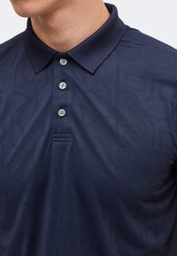 Homme portant un polo bleu marine avec un motif subtil du logo Calvin Klein et trois boutons blancs sur la patte de boutonnage.