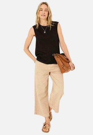 Schwarzes ärmelloses Oberteil, beige weit geschnittene Hose, braune Schultertasche und gemusterte Sandalen, Model steht vor einem weißen Hintergrund.