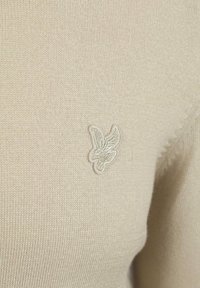 Beige gebreide trui met een subtiele geborduurde logo. Het materiaal is soepel, met fijne ribbels en een getailleerde vorm.