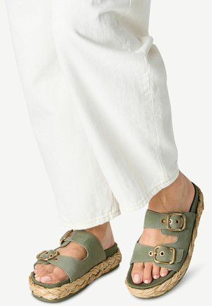 Vihreät mokkanahka-sandaalit, joissa on kaksi säädettävää solkea, kudottu juutti-välikerros ja teksturoitu kumipohja, yhdistettynä valkoisiin housuihin.