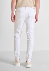 Jeans de mezclilla blanca con un corte slim, bolsillos traseros y una textura suave y lisa; combinados con zapatillas beiges sobre una superficie clara.