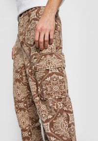 Brune cargo bukser med intrikate beige mønstre, lavet af stof med en tekstureret overflade. Har sidelommer og en afslappet pasform.