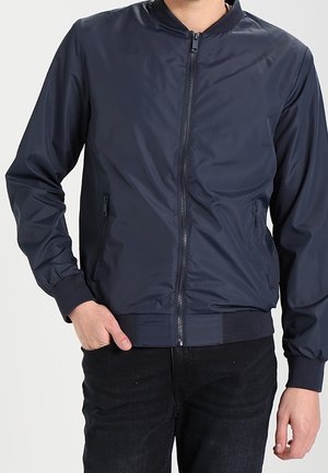 Mann trägt eine marineblaue Bomberjacke mit Reißverschluss, gerippten Bündchen und Saum, kombiniert mit schwarzer Hose, eine Hand in der Hosentasche.