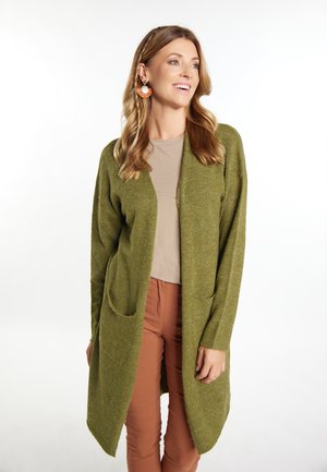 Olijfgroene gebreide cardigan met een open voorkant, twee voorzakken en geribde manchetten, gedragen met een beige top en een bruine broek.