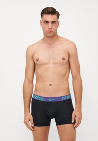 Črne bokserice z violettno in zeleno Nike logotip obrobo, z oprijetim krojem in gladko teksturo, model stoji z nevtralnim izrazom.