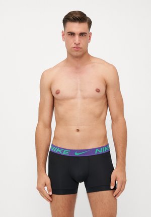 Sorte boxer shorts med en lilla og grøn Nike-logo elastik i taljen, som har en tætsiddende pasform og glat tekstur. Modellen står med et neutralt udtryk.