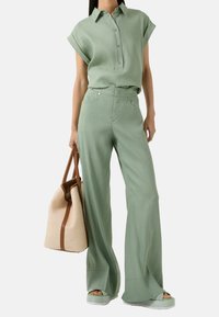 Donna che indossa una camicia button-down a maniche corte verde chiaro e pantaloni ampi coordinati, mentre tiene una borsa a tote beige con manici marroni.