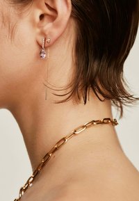 Los pendientes de cadena dorada presentan una gota de piedra preciosa púrpura y una barra delgada y colgante, resaltando detalles intrincados y texturas contrastantes.