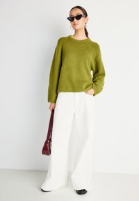 Ženska v velikem olivno-zelenem puloverju, širokih belih hlačah, črnih sončnih očalih, uhani v obliki obroča, drži bordo torbico proti enobarvni podlagi.