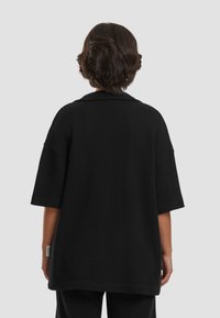 Svart ribbestrikket oversized skjorte med korte ermer og notched krage. Stoffet ser ut til å være teksturert. Lappen er synlig på sidesømmen.