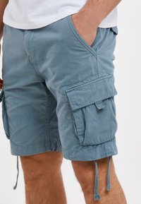 Pantaloni cargo azzurri chiari con numerose tasche, orli con coulisse e un tessuto morbido e texturizzato. Cuciture a vista e una vestibilità comoda.
