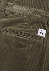 Olijfgroene corduroy broek met een gestructureerd ribbelpatroon, voorzien van een achterzak en een wit label met "M 73" in het zwart.