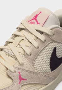 Close-up van een beige en crèmekleurige Nike sneaker met een roze Jumpman-logo op de tong en zijkant, gemaakt van mesh en suède materialen en met crèmekleurige veters.