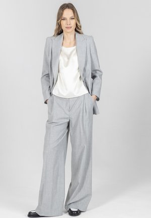 Kobieta w jasnoszarym garniturze z szerokimi nogawkami, z dopasowaną marynarką i białą satynową bluzką, stoi z rękoma w kieszeniach na jednolitym tle.