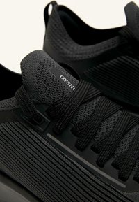 Czarne buty sportowe z teksturowaną, dzianą cholewką i płaskimi sznurowadłami. Boki mają elegancki design z subtelnym logo i paskowanymi detalami.