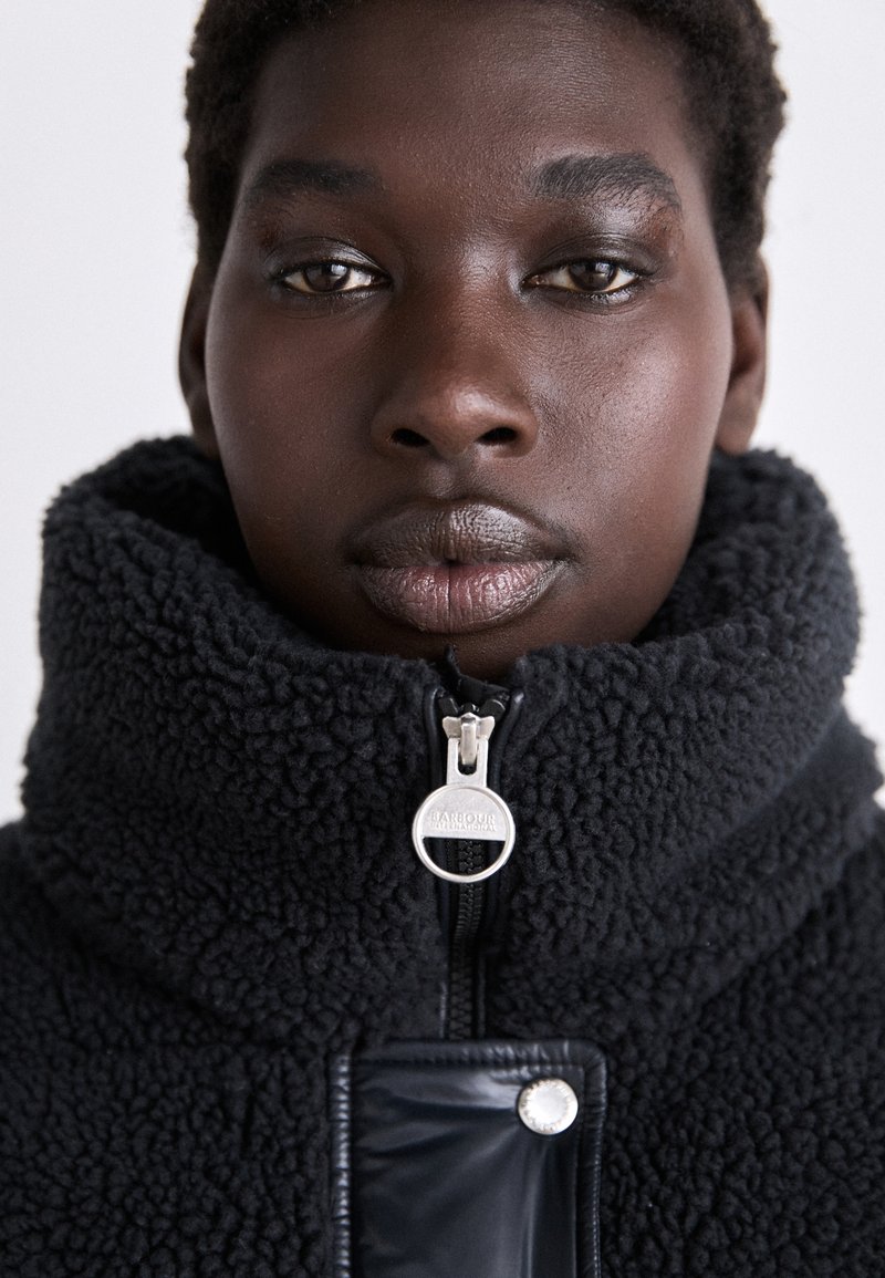 Giacca in pile nera con colletto alto, caratterizzata da una zip argentata e dettagli in pannello nero lucido. Il tessuto texturizzato contrasta con le finiture lisce.