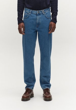 Muž v modrých džínách přímo střižených nohavicích a hnědých šněrovacích botách, stojící na světlém jednobarevném pozadí.