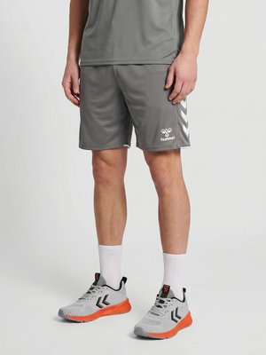 Mann trägt graue Sportshorts und passendes Shirt, weiße Sportsocken und graue sowie orange Sportschuhe, steht vor einfachem Hintergrund.