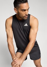 Camiseta deportiva sin mangas negra con logo blanco, combinada con pantalones cortos oscuros. Tejido suave, diseño ajustado que muestra brazos musculosos.