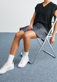 Svart stickad ärmlös topp med V-ringning, grå veckad kjol, vita strumpor och vita sneakers; sitter på en kromad hopfällbar stol.