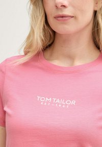 Rožinė medvilninė marškinė su apvalia apykakle ir trumpomis rankovėmis. Išskirtinė balta logotipo spausdinimo "TOM TAILOR EST-1962" detalė priekyje.
