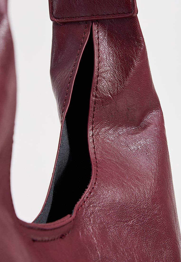 Bolso de cuero burdeos con una textura suave, apertura curva y detalles de costura sutiles. El interior es negro, resaltando la forma del bolso.