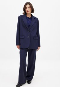 Navy blue tailored blazer with notch lapels, two front pockets, and single button closure, paired with wide-leg trousers and polished shoes.