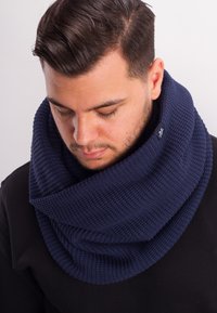 Navy blue knit infinity scarf featuring a textured pattern and a logo accent, draping around the neck and shoulders.