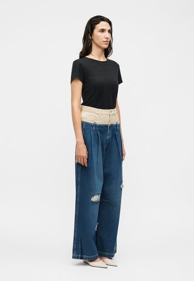 Femme debout portant un t-shirt noir à manches courtes, un jean large taille haute bicolore avec une ceinture beige, des détails déchirés et des chaussures blanches pointues.