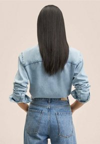 Chemise en denim bleu clair avec manches retroussées, vue de dos. Associée à un jean taille haute bleu clair, montrant deux poches arrière.