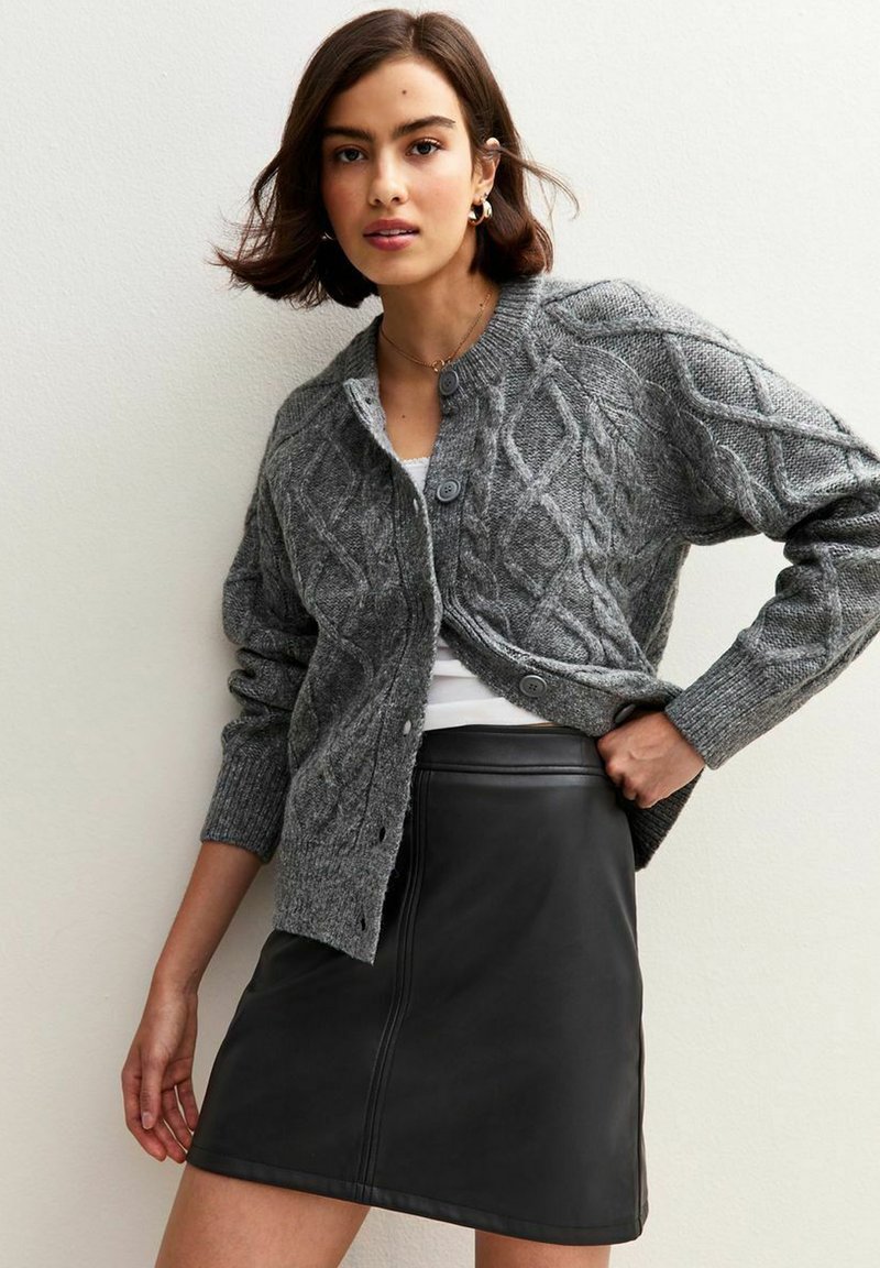 Grijze, gebreide cardigan met een kabelpatroon, voorzien van een ronde halslijn en knoopsluiting, gedragen over een witte top en een zwarte leren rok.