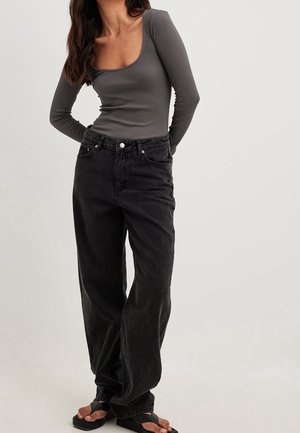 Mujer con una camiseta gris ajustada de manga larga y escote redondo, jeans negros sueltos y sandalias negras, de pie con las manos detrás de la espalda.