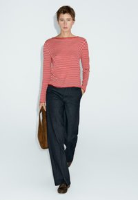 Vrouw die loopt in een rood-wit gestreept lange mouwen top, met een brede broek in het donker, bruine instappers draagend en een bruine suède tas meedragend.