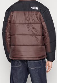 Osoba w brązowej i czarnej kurtce puchowej z logo The North Face na górnej części pleców, w połączeniu z ciemnoniebieskimi dżinsami z białym przeszyciem.