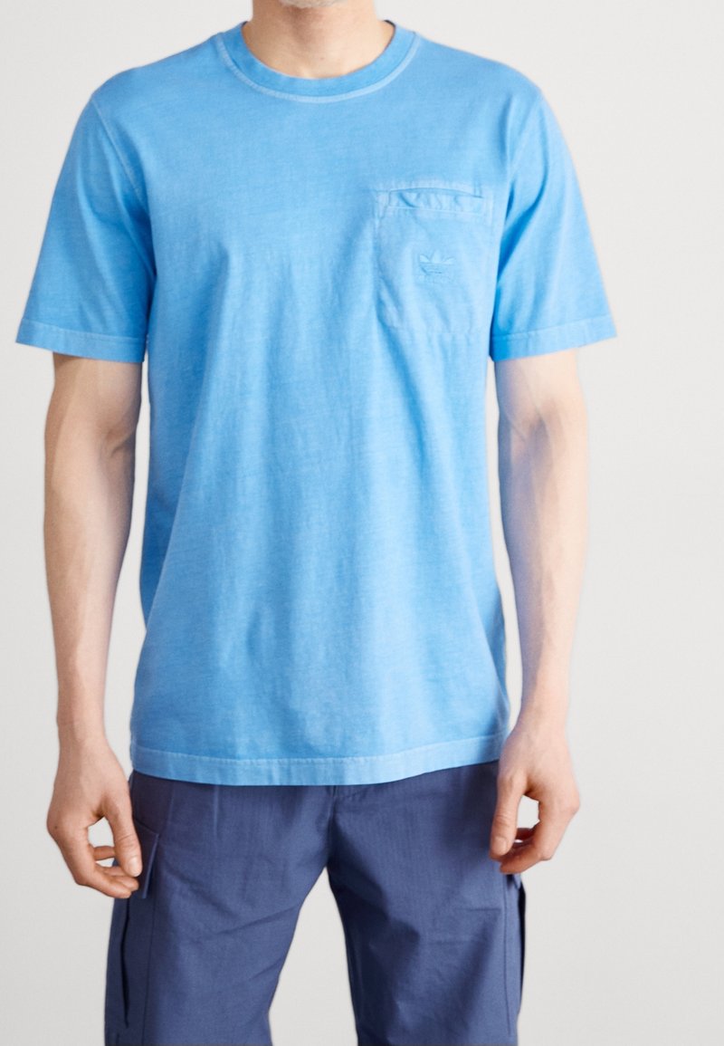Světle modré bavlněné tričko s krátkými rukávy a kulatým výstřihem. Obsahuje malou kapsu na levé části hrudi s nenápadným logem.