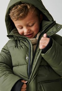 Giacca invernale imbottita verde con cappuccio, dotata di chiusura con cerniera e bottoni, polsini elastici e un esterno in nylon liscio.