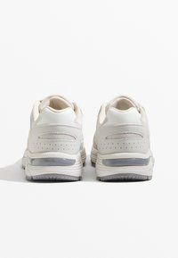 Jasne sportowe buty wykonane z materiałów siatkowych i skórzanych, o zaokrąglonym kształcie, z wyraźnym amortyzatorem pięty oraz perforowanymi detalami po bokach.